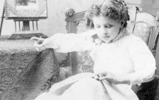 A monochrome archive photograph shows Helen Keller, around the age of eight, seated in a high barked armchair, next to a cloth-covered table, her legs resting upon a footstool. Clothed in a knee-length white dress and ankle boots, she reads with her left hand and points with her right hand out of shot.