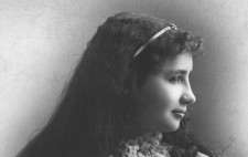 A monochrome archive studio portrait shows sitting turned to the right of the frame. She wears a white dress, with a corsage, and ruched at the neck. Her long dark hair flows over her shoulders and she smiles.