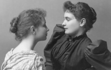 A monochrome archive photograph shows a teenage Helen sitting on a chaise facing a young Annie. Both are dressed elegantly, their hair gathered into buns, and Helen in a white dress with a draped floral shawl, Annie in black with mutton leg sleeves.