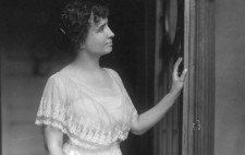 A monochrome archive photograph shows Helen, in her 20s, standing in an ornately carved doorway wearing a lavish floor-length frock dressed in layers of white lace. Her her left hand rest lightly on the door and she faces towards the right of the frame, her face pensive.