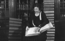 A monochrome archive photograph shows Helen dressed smartly in a black dress with pearls around her neck, sitting in a library, where print books behind glass fill oak shelves. On her lap rests a large Braille book. Next to her, stacks of Braille books tower twenty-high.