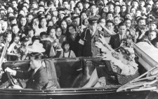 Helen and Annie sit in an open top car. In the background a deep crowd of people can be seen smiling and jostling to get a look at her. Helen has her arms outstretched, waving at the crowd.