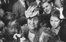 A monochrome archive image shows Helen, in her sixties, amongst children dressed in school uniforms, who crowd in to get close to her. She has her arms around three of the children.