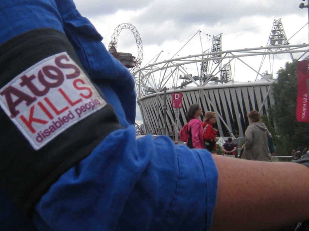 Worn over Liz Crow's blue shirt is a close-up of the Atos protest armband, showing parts of the slogan 'Atos kills disabled people' in purple and red on white. In the background is the Olympic stadium and a banner saying 'London 2012'.