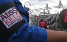 Worn over Liz Crow's blue shirt is a close-up of the Atos protest armband, showing parts of the slogan 'Atos kills disabled people' in purple and red on white. In the background is the Olympic stadium and a banner saying 'London 2012'.