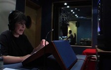 Director Liz Crow sits in a small studio recording audio description. She wears earphones and speaks into a microphone. On a small stand in front of her is a script and she is looking up to a monitor which plays the film. Beyond the studio window a sound editor sits at a mix desk.