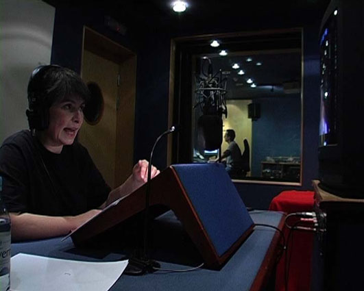 Director Liz Crow sits in a small studio recording audio description. She wears earphones and speaks into a microphone. On a small stand in front of her is a script and she is looking up to a monitor which plays the film. Beyond the studio window a sound editor sits at a mix desk.