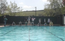 Seven boys in swimming costumes sit along the edge of a swimming pool. Walter sits with feet in the water, but still wearing a dressing gown. Coach stands in the background, wearing a stripy blazer and cream hat. Next to him is Rachel, holding notes and a walkie-talkie. To the left of the pool stands the sign language interpreter.