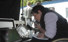 Under a black awning, Director Liz Crow sits on her wheelchair, looking intently at a monitor and holding a walkie-talkie ready to speak. Behind her in bright sunshine is the camera crew.