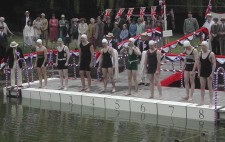 Eight swimmers line up along a pontoon festooned with patriotic red, white and blue, preparing to dive into lake water. The marshall, in navy blazer and straw boater, stands to one side. Behind the swimmers, supporters crowd the grassy banks.