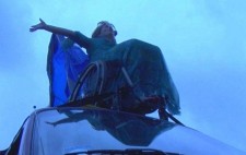 Against a half-light blue sky, a woman sits on her wheelchair, high up on top of a van roof. In a long dress, her arms thrown wide, blue fabric draped behind her, she tips back her head and whoops her joy.