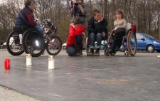 A group of five people from the production team gather around a weathered bronze memorial plaque set into pavement. Behind them are parked cars and the parkland trees of Tiergartenstrasse. Three votive candles have been set on the memorial.