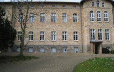 A block paving driveway with mown lawns either side leads to a four storey yellow-brown brick institution, a row of half-basement windows running the length of the building.