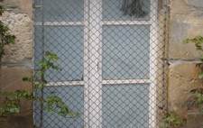 A close up of the outside of one of a lower ground floor window, whited out and with a metal grill over it. On the ground are scatted wood chippings and leaves from nearby planting frame the window.