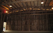 A photograph shows a cavernous barn-like wooden structure dwarfs a door open to the outside where people peer in. 