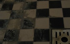 A close up of a black and white tiled floor. Across the floor is a track of water stain and limescale, which passes by a small square drainage grid.