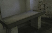 A small room with rough limewashed stone walls and rough stone floor, a small window up high. In the centre is a grey stone table with raised sides, a drainage pipe at one end which runs to the floor.