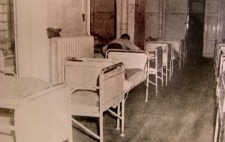 An archive photograph shows an old-fashioned hospital ward tightly packed with rows of empty metal-framed beds. A member of staff makes up one of the beds.