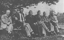 An archive photograph shows six men, ranging in age from thirties to fifties, wearing suits and ties, sitting in a row on benches under a tree in a park. Turned to the camera, their faces are neutral.