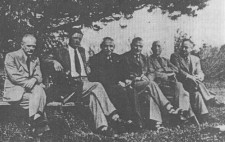 An archive photograph shows six men, ranging in age from thirties to fifties, wearing suits and ties, sitting in a row on benches under a tree in a park. Turned to the camera, their faces are neutral.