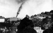 A gothic institutional building built high above a small town, black smoke streaming from its chimney.