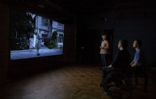 In a black box space, an audience of three watches the historical drama on a large screen. The shot of the institution forecourt appears suspended in pitch dark.