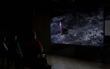 An audience watches the drama on the large screen suspended in pitch dark space. Filmed from above, a woman, dressed in a shabby pink dress and cardigan, sweeps a dark path across a large swathe of gravel.