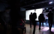 In a dark gallery space, the film on the first screen has ended and the screen shows bright white. Audience members move around the side of the screen into the space behind.