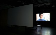 In the darkened gallery beyond the first screen, Lou is projected in close up, shown in a domestic setting, greenery visible in the window behind her.