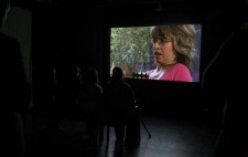 An audience sits in front of the second screen, watching Sophie projected in close-up, shown in a domestic setting, greenery visible in the window behind her.