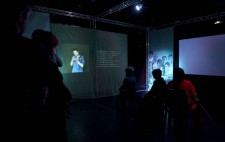 A group of people sit and stand in the dark of the Resistance installation surrounded by green banners. On a plain green area of banner, a projected image shows a woman signing. To her right the words of the speaker are projected onto the banner wall. In the distance is a spotlit banner, a montage of faces looking out to the viewer.