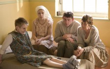 A group of four inmates gather round together. Three perch in a row on a bed, leaning towards another who sits on a neighbouring bed with her legs stretched out in front of her and arms extended behind her for support. All of them gaze far distance, their expressions grim.