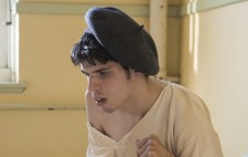 A close up of one of the inmates. He sits on the edge of a bed, wearing an ill-fitting white fabric top and a black beret, his face written with anxiety.