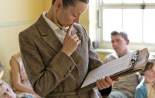 An office, dressed in a 1930s grey tweed suit, stands in the middle of the ward examining a typed list on her  clipboard. Behind her, inmates sit or lie on dormitory beds.