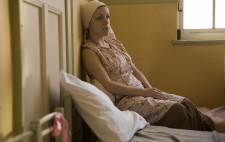 An inmate sits on a bed and rests up against the wall behind her, a window to her right. She wears white headscarf and floral dress and gazes far distance.