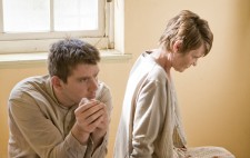 Two of the inmate extras sit on a bed between takes, both gazing far distance. One, a man dressed in a grey shirt and ripped green trousers, sits facing the camera. The other, a woman dressed in a grey cardigan and a checkered skirt, sits with her back to him.