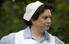 The Nurse stands with her hands on her hips, dressed in a pale blue nurse's outfit and white apron, a small Nazi swastika badge on her lapel.