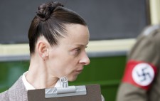 During the bus loading scene, a young woman in a grey 1930s suit, turns towards the right of the frame, her hair pulled tight into a bun. She clutches a clipboard and pencil to her chest. In the background stands a man wearing a Nazi swastika armband.