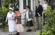 Production still. A nurse leads five of the inmates out of the institution. One is on crutches.