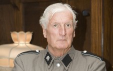 A man in a grey military style outfit and wearing a Nazi swastika armband stands, arms crossed, looking at the camera.