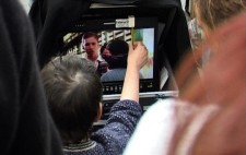 On a monitor is a close up of Walter and Gloria. Liz holds a pen up to the monitor quarter of the way in from the right of the screen to demonstrate to the crew the space that will be occupied by the interpreter.