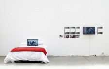 In a white-walled gallery, is a double bed with a white quilt and a throw of red fabric. Leaning against the pillows is a video display screening Reflections from the Bed. On the wall behind are images from...