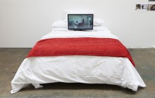 In a white-walled gallery, is a double bed with a white quilt and a throw of red fabric. Leaning against the pillows is a video display screening Reflections from the Bed.