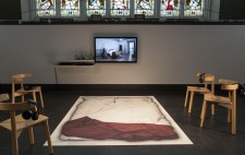 A large mat displaying a life-sized photograph of Liz's empty double bed, white bedding and a red fabric throw, lies flat on the black floor of the stage. Grouped around it are four chairs, each with a headset looped over its back. At the pillow end of the   mat, mounted on the wall, is a screen showing the film Reflections from the Bed.