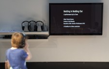 A child looks up at a wall-mounted TV monitor which displays the credits for the film.To the left is a row of three cordless headsets.