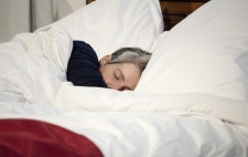 A close up of Liz lying amongst white sheets. She wears a blue top, and a throw of red fabric drapes across the quilt.