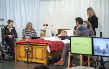 Liz Crow lies on the stage in her bed, with four people seated around it, holding microphones. A sign language interpreter stands to the side of the bed. Stage right are two monitors.