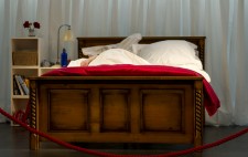 The dark pine bed photographed from the footboard, a wall of white drapes behind. Liz is almost hidden beneath the covers. Next to the bed is a white unit of cubes with books, a lamp, wicker baskets and a small vase of flowers. A red rope loops from left to right forming a barrier between the viewer and the bed.