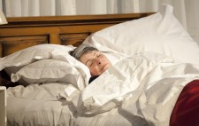 A close up as Liz lies asleep in bed, her head sunk into pillows and a quilt drawn high to her chin.