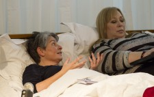 A close up of Liz Crow and Dawn Willis, reclined next to each other on the bed. Liz looks over to Dawn and gesticulates, as Dawn types.